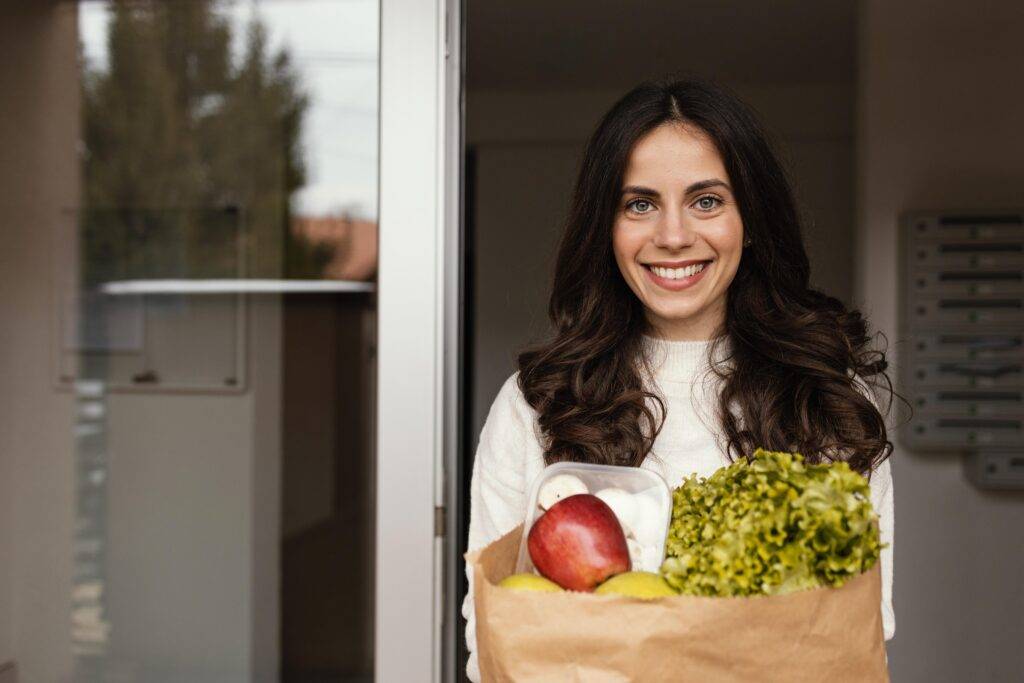 A smiling personal shopper placing a grocery bag on a client’s kitchen counter, with the two chatting warmly. The image conveys trust, familiarity, and ongoing service.