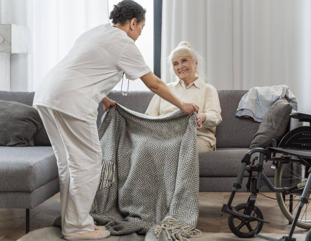 A caregiver helping an elderly person in a kitchen, with fresh groceries and household supplies on the counter. The image conveys care, reliability, and support for daily living needs.