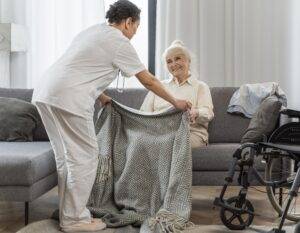 A caregiver helping an elderly person in a kitchen, with fresh groceries and household supplies on the counter. The image conveys care, reliability, and support for daily living needs.