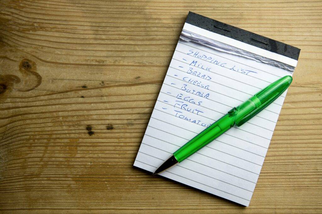A handwritten grocery list on a notepad beside fresh produce and a cup of coffee. The image suggests thoughtful planning and practical shopping.